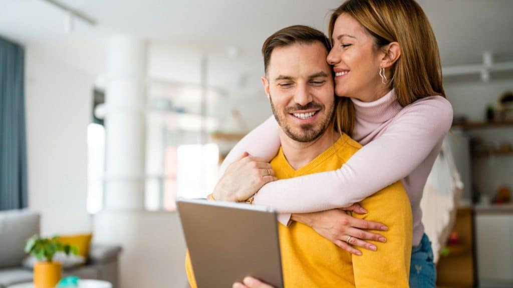 A woman in a pink top hugs a smiling man from behind, who holds a tablet.