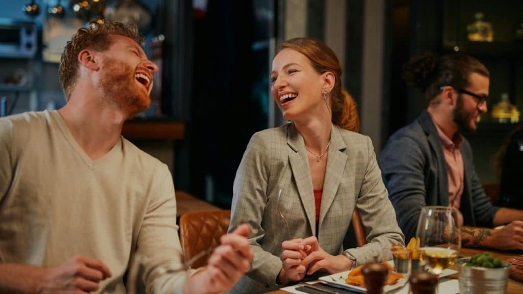 A red-headed man and a woman in a blazer laugh together at a table in a restaurant.