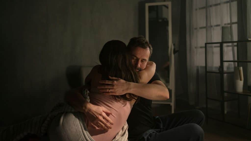 A man embraces a woman, both looking distressed, in a dimly lit room.