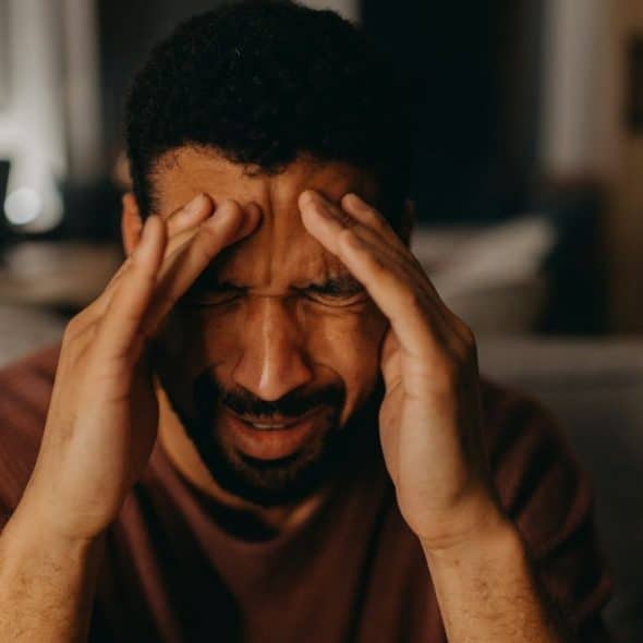 A distressed man with his eyes closed holds his hands to his temples, grimacing.