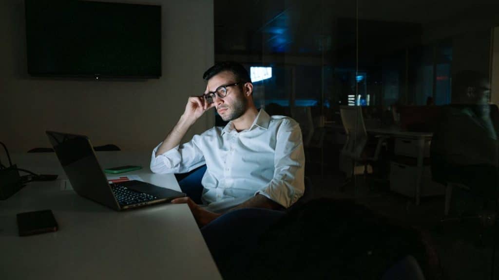 A man in glasses and a white shirt looks tired while working on a laptop in a dimly lit office.