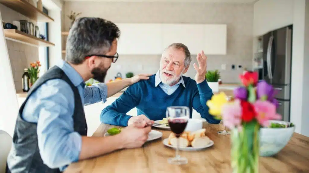 A younger man places a hand on the shoulder of an older man, who is gesturing while seated at a table.