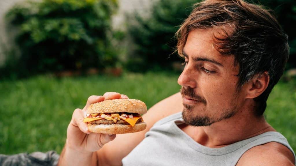 A man with a beard and messy hair lies on grass, holding a burger.