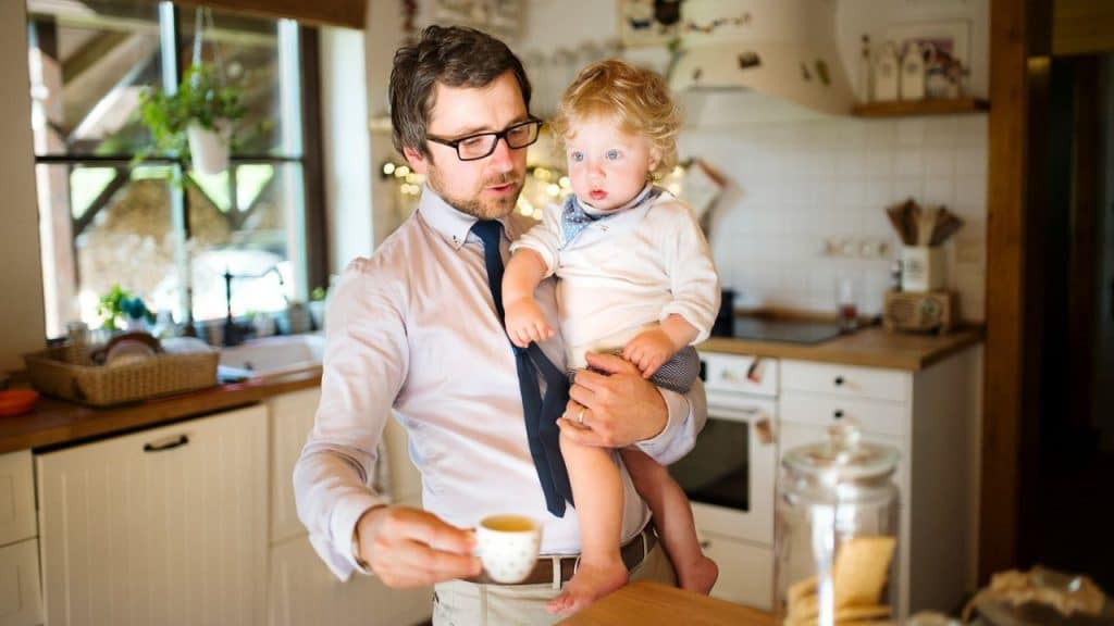 A man in a dress shirt and tie holds a baby and a cup of coffee in a kitchen.