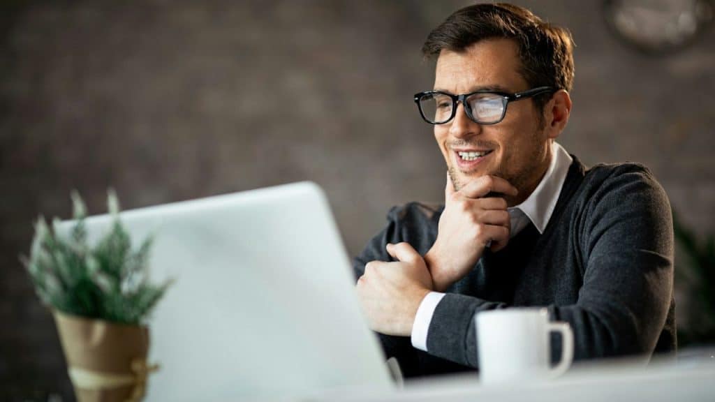 A smiling man with glasses looks at a laptop, resting his chin on his hand.