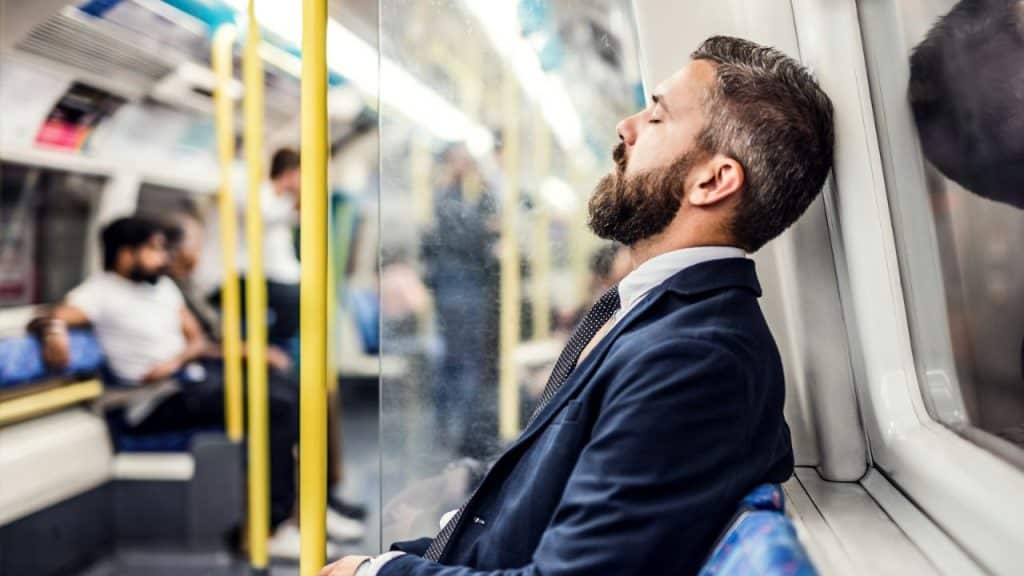 A bearded man in a suit sleeps with his head against a window on a train.