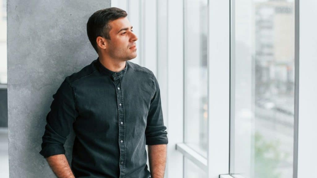A man in a dark shirt stands by a window, looking out thoughtfully.