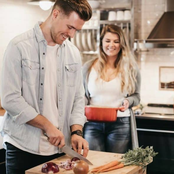 A man smiles as he chops an onion on a cutting board, while a woman smiles behind him.