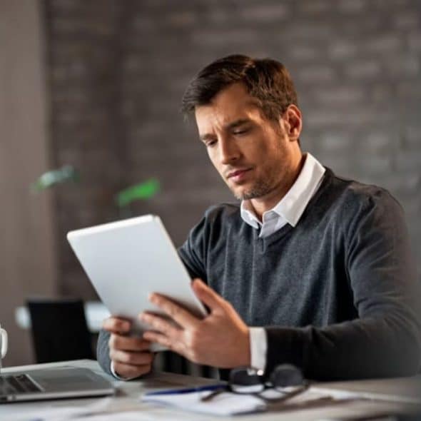 Man holding a tablet looking thoughtfully across a shared workspace.