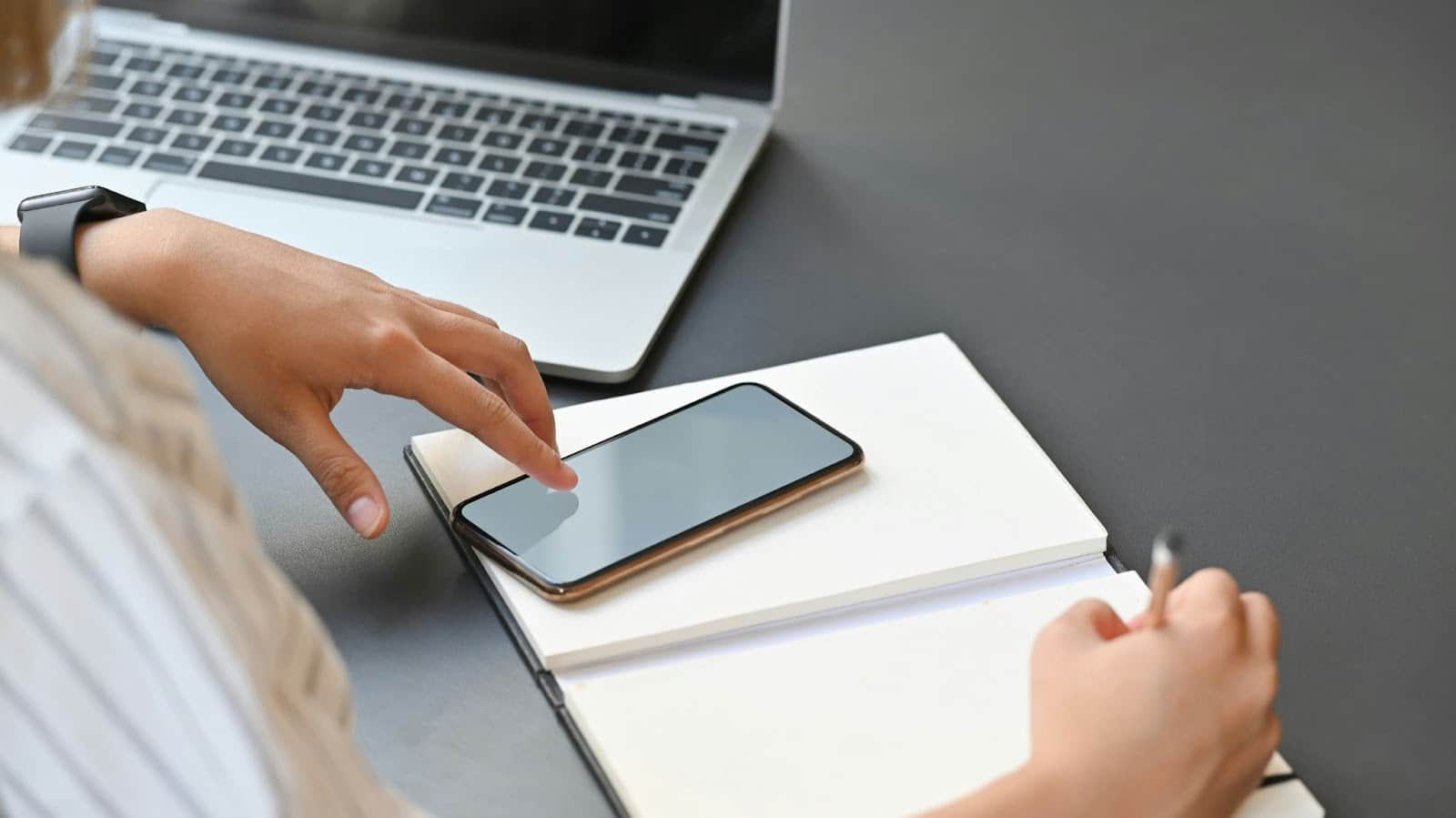 A man writing on a book while checking his phone.