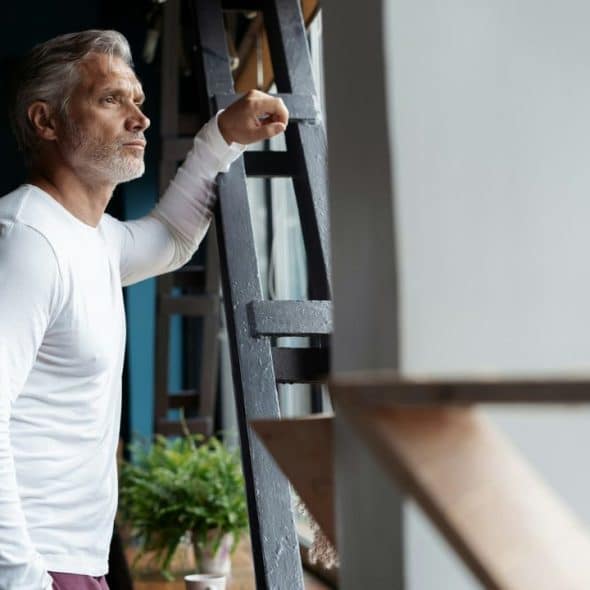 A mature man with gray hair and a white long-sleeved shirt looks out a window, resting his arm on a metal structure.