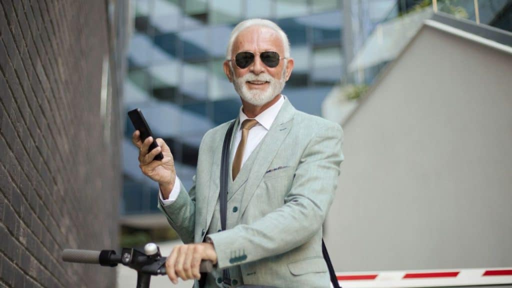 A stylish elderly man in a suit and sunglasses smiles while holding a phone and scooter.