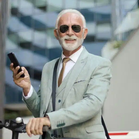 A stylish elderly man in a suit and sunglasses smiles while holding a phone and scooter.