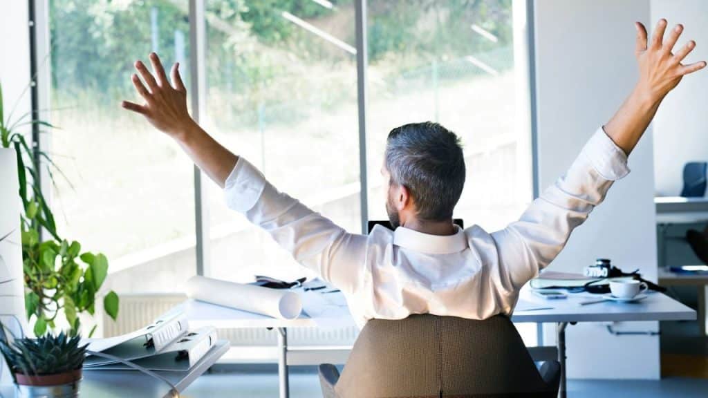A man with graying hair sits at a desk facing a window, stretching his arms upwards.