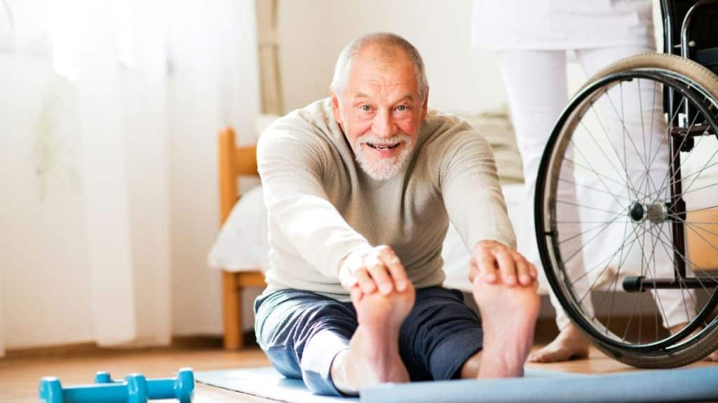 A smiling elderly man stretches forward on a mat, touching his toes, with a wheelchair nearby.