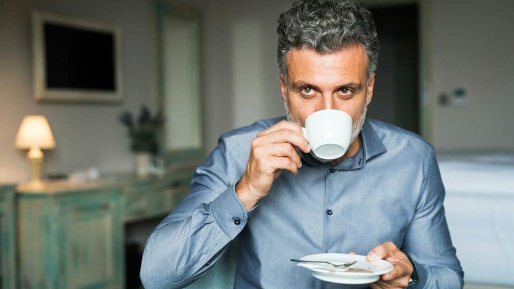 A grey-haired man with a beard sips from a white mug, looking directly at the viewer.