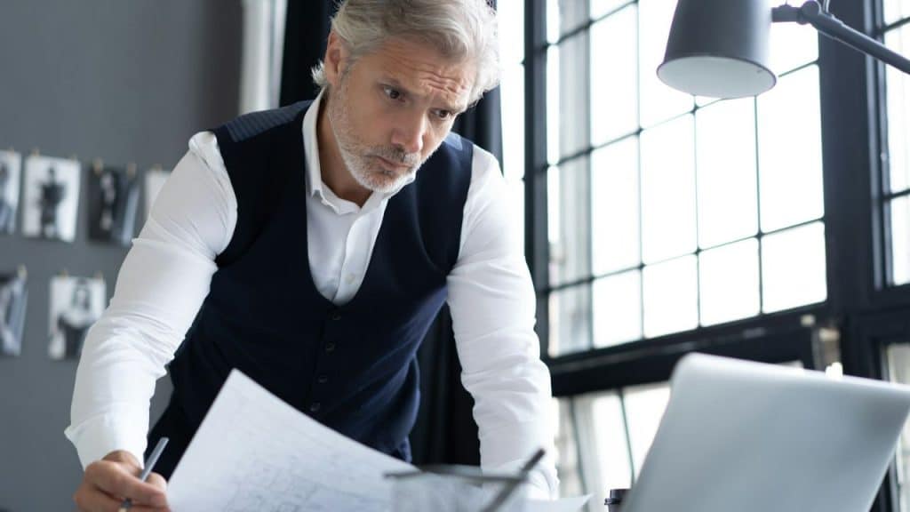 A focused, grey-haired man in a white shirt and dark vest reviews a document.