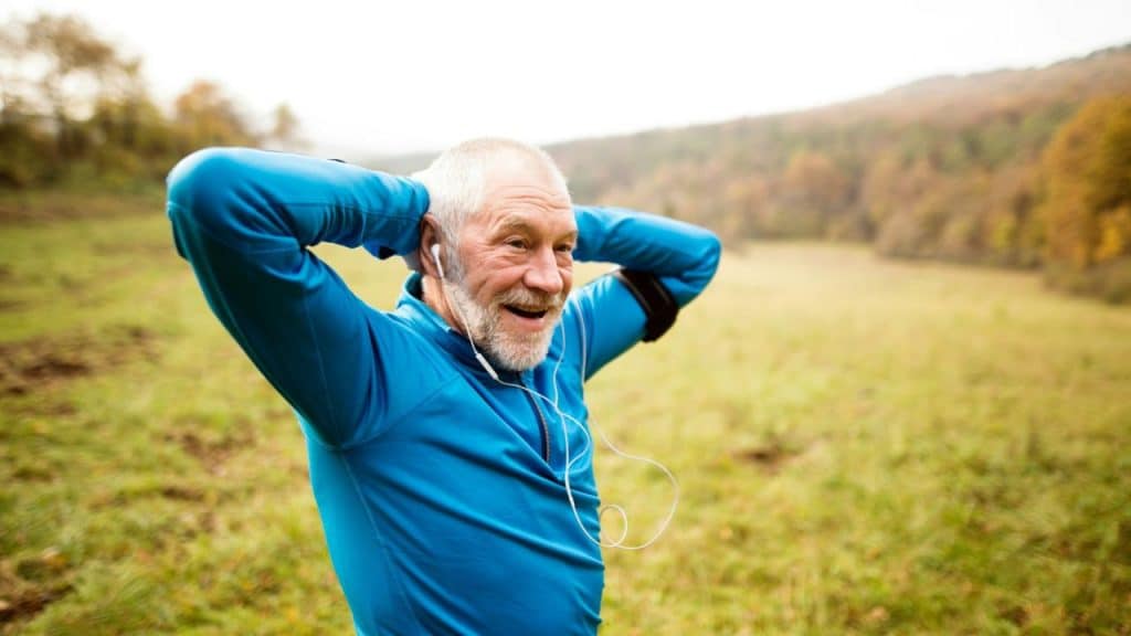 A smiling elderly man in a blue long-sleeved shirt and earbuds stretches outdoors.