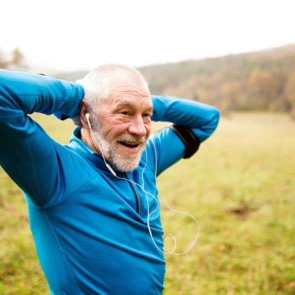 A smiling elderly man in a blue long-sleeved shirt and earbuds stretches outdoors.