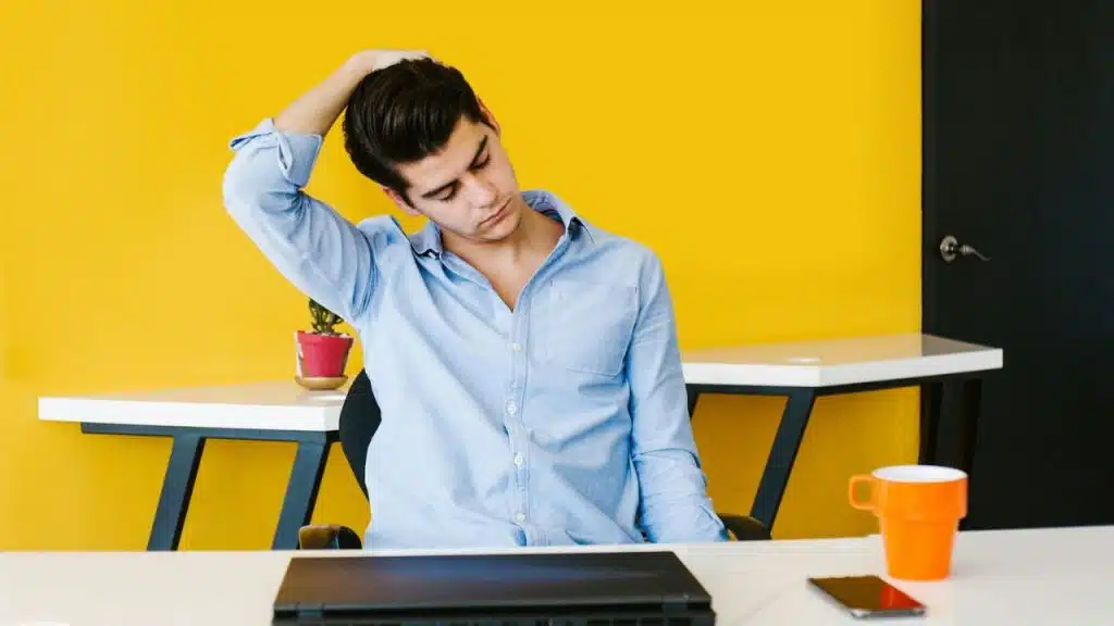 A man in a blue shirt stretches his neck while sitting at a desk with a laptop.