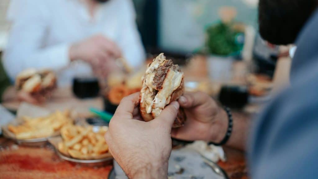 A person holds a large, half-eaten burger, with fries and other diners blurred in the background.