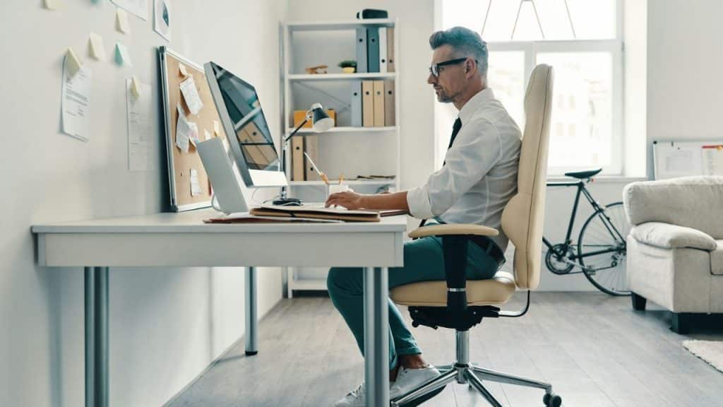 A grey-haired man in a white shirt and teal pants works at a computer desk.