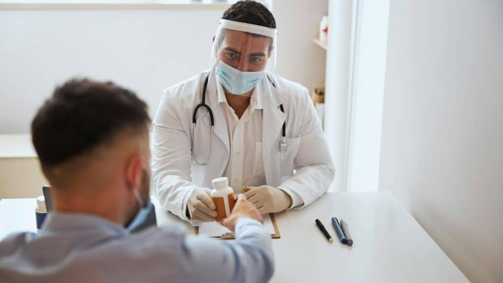 A doctor in a mask and face shield hands a bottle to a patient across a white desk.