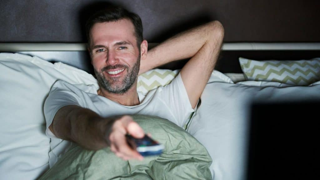 A smiling man with a beard reclines in bed, holding a TV remote control.