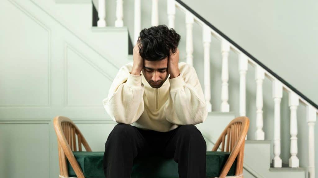 A man with dark, curly hair sits, holding his head in his hands, looking distressed.