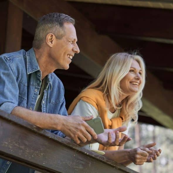 A smiling mature couple stands on a balcony, looking outwards.