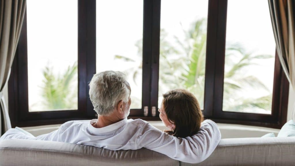 An older couple from behind, looking out a large window at palm trees.