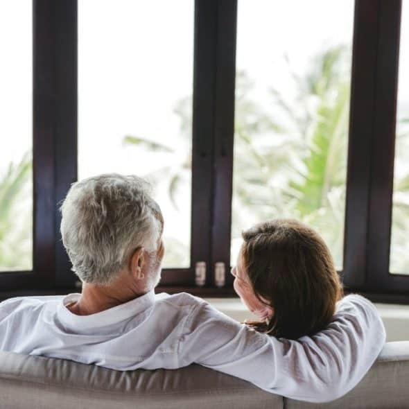 An older couple from behind, looking out a large window at palm trees.