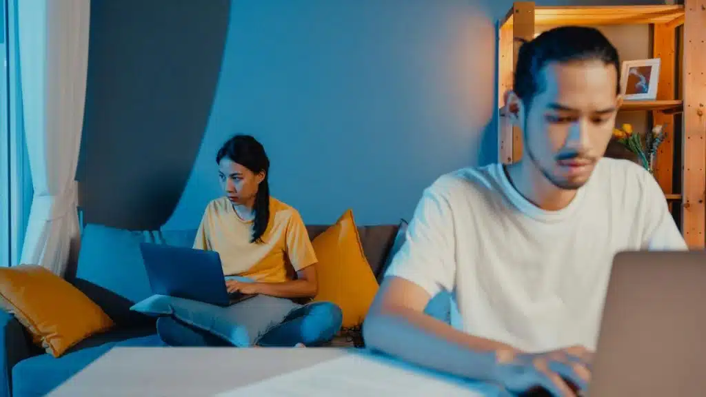 A man and a woman are in a living room, each focused on their laptops.