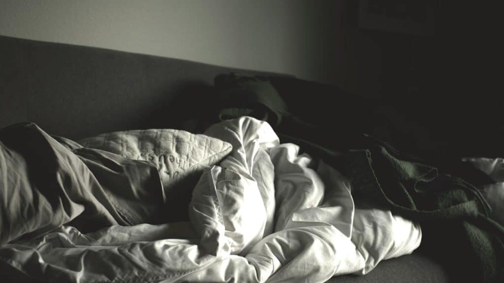 A gray couch covered in crumpled white sheets and a dark blanket.
