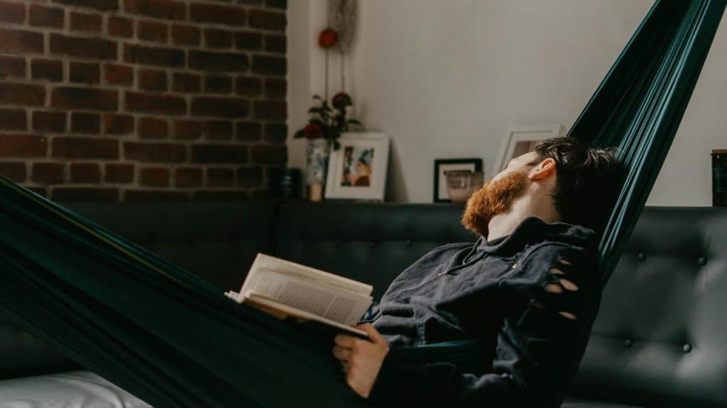 A bearded man reading a book while reclining in an indoor hammock.