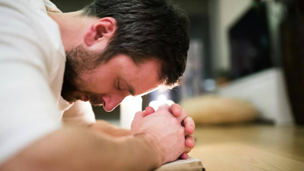 A man leaning forward in prayer with his hands clasped over an open book.
