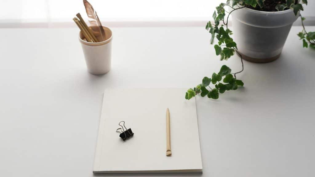 A minimalist workspace with a notebook, pencil, binder clip, cup of pens, and a potted plant on a white desk.
