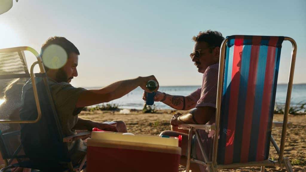An image of two men relaxing on beach chairs by the shore, sharing a drink in the evening sunlight.