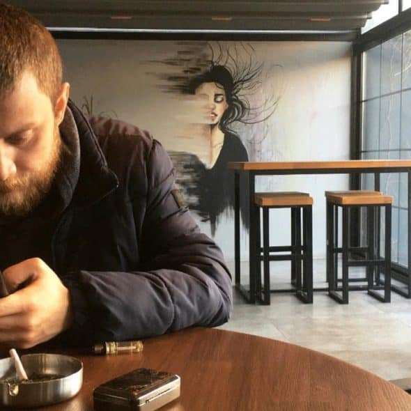 A bearded man at a café table scrolling on his phone, ashtray and vape in front, with a mural and windows behind.