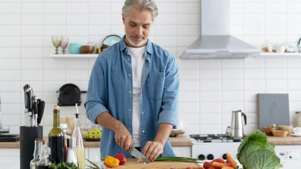 A grey-haired man in a denim shirt chops vegetables on a cutting board in a kitchen.