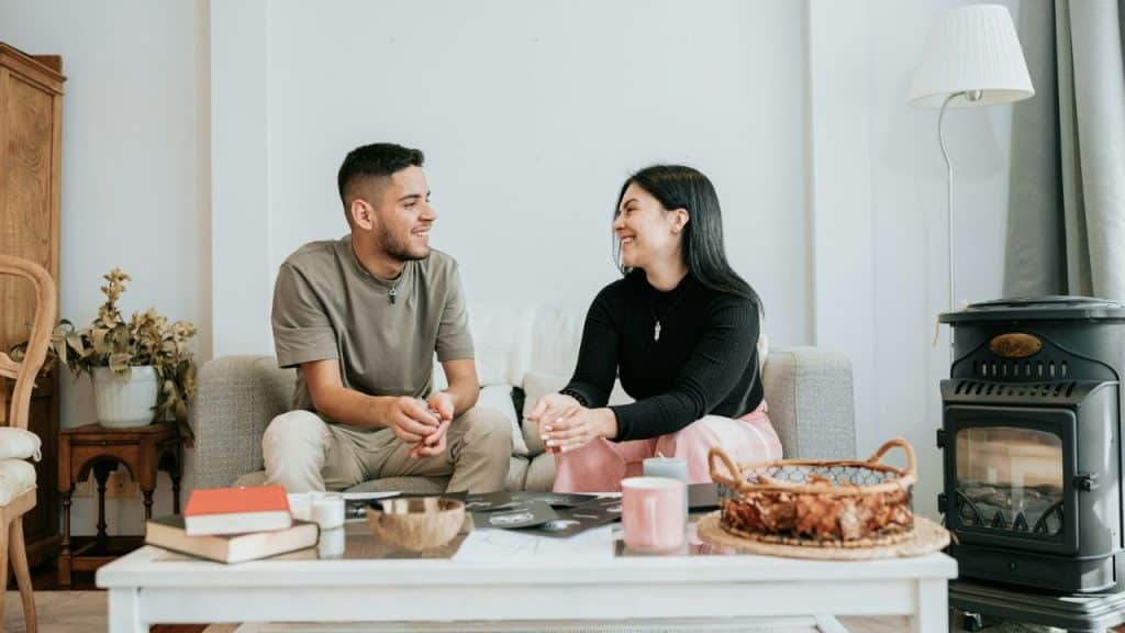 A smiling man and woman sit on a couch, facing each other and talking.