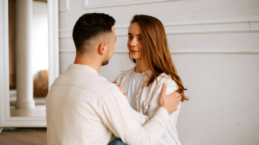 A man in a white shirt faces a woman with long brown hair, who looks at him while he holds her arms.