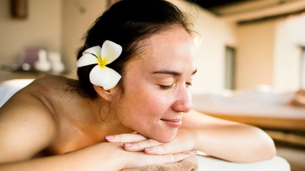 A woman with a white flower in her hair rests her head on her hands, smiling with eyes closed.