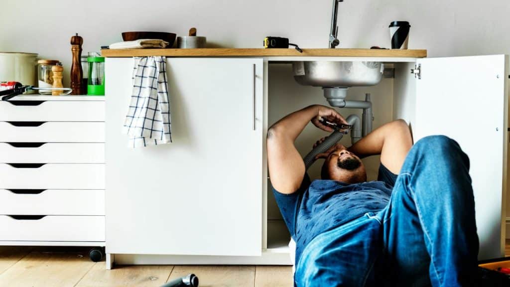 A man lies on his back under a kitchen sink, fixing pipes.