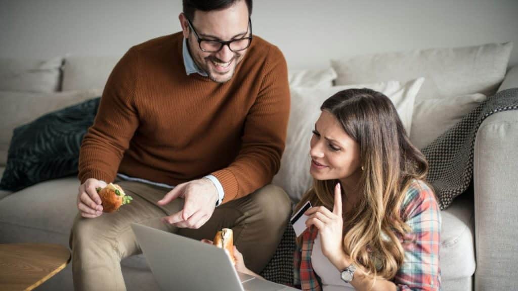 A smiling man with glasses holds a sandwich while a woman next to him holds a credit card and looks at a laptop.