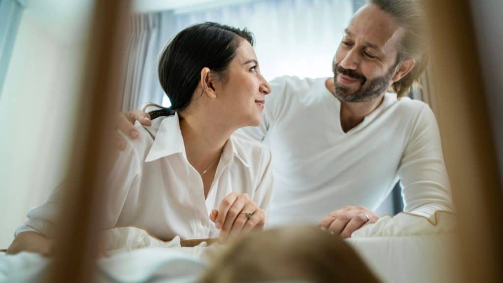 A smiling man and woman gaze at each other over a baby's crib.