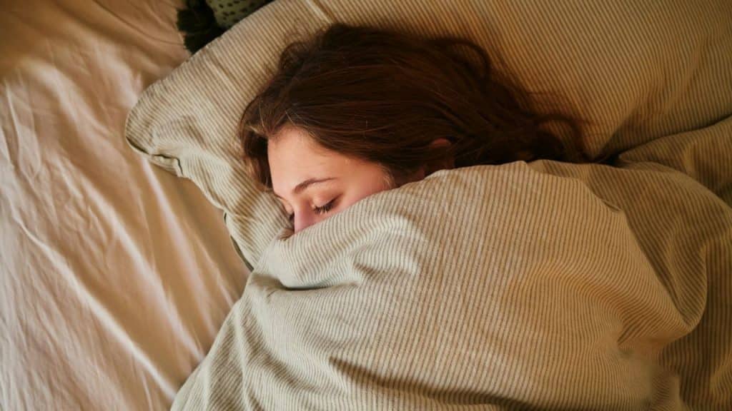 A person with long brown hair sleeps soundly under a striped blanket.
