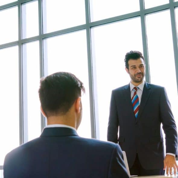 A man in a suit smiles and looks at three other men in suits in a room with large windows.