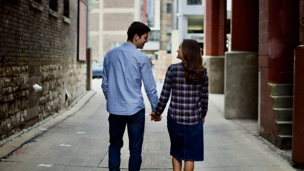 A man in a blue shirt and a woman in a plaid shirt and denim skirt walk hand-in-hand, looking at each other.