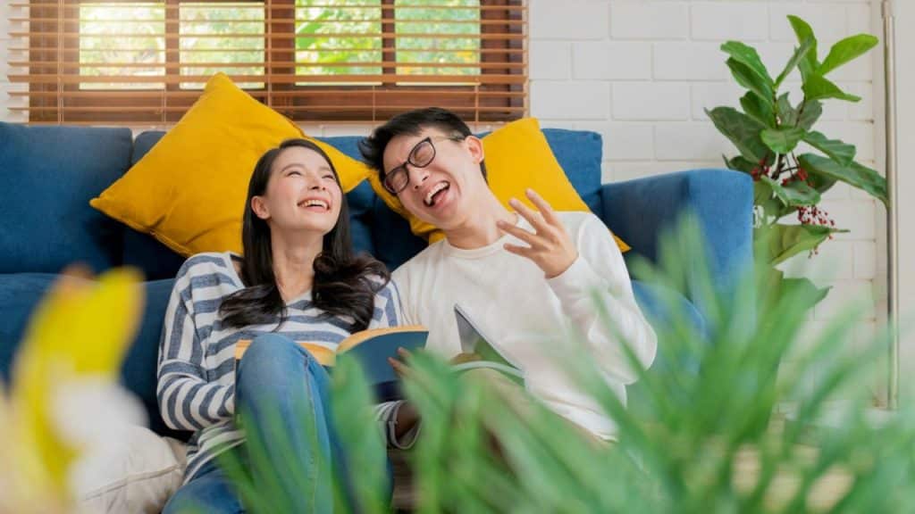 A joyful couple, sitting by a blue couch, laughs while holding books.